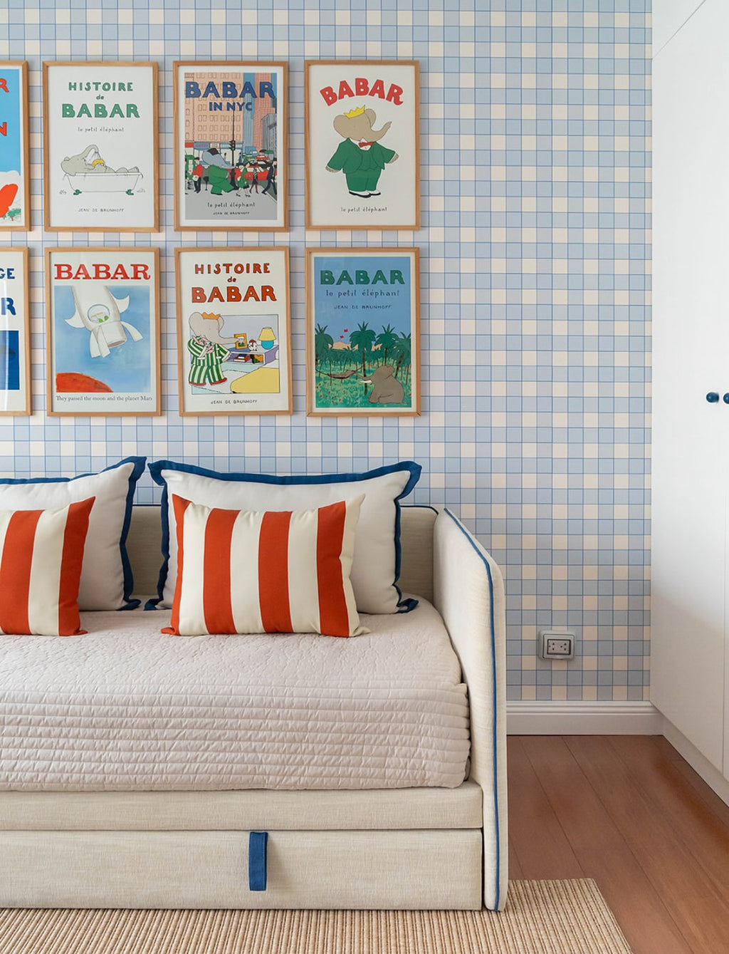 Papier peint Jack à motif damier bleu et blanc, décorant le mur derrière un lit banquette avec des coussins rayés rouge et blanc et des affiches vintage Babar encadrées.