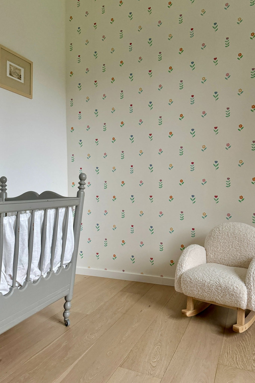 Échantillon du papier peint Juliette à motif de petites fleurs, décorant une chambre de bébé avec un lit à barreaux gris vintage et un petit fauteuil à bascule en tissu bouclette blanc.