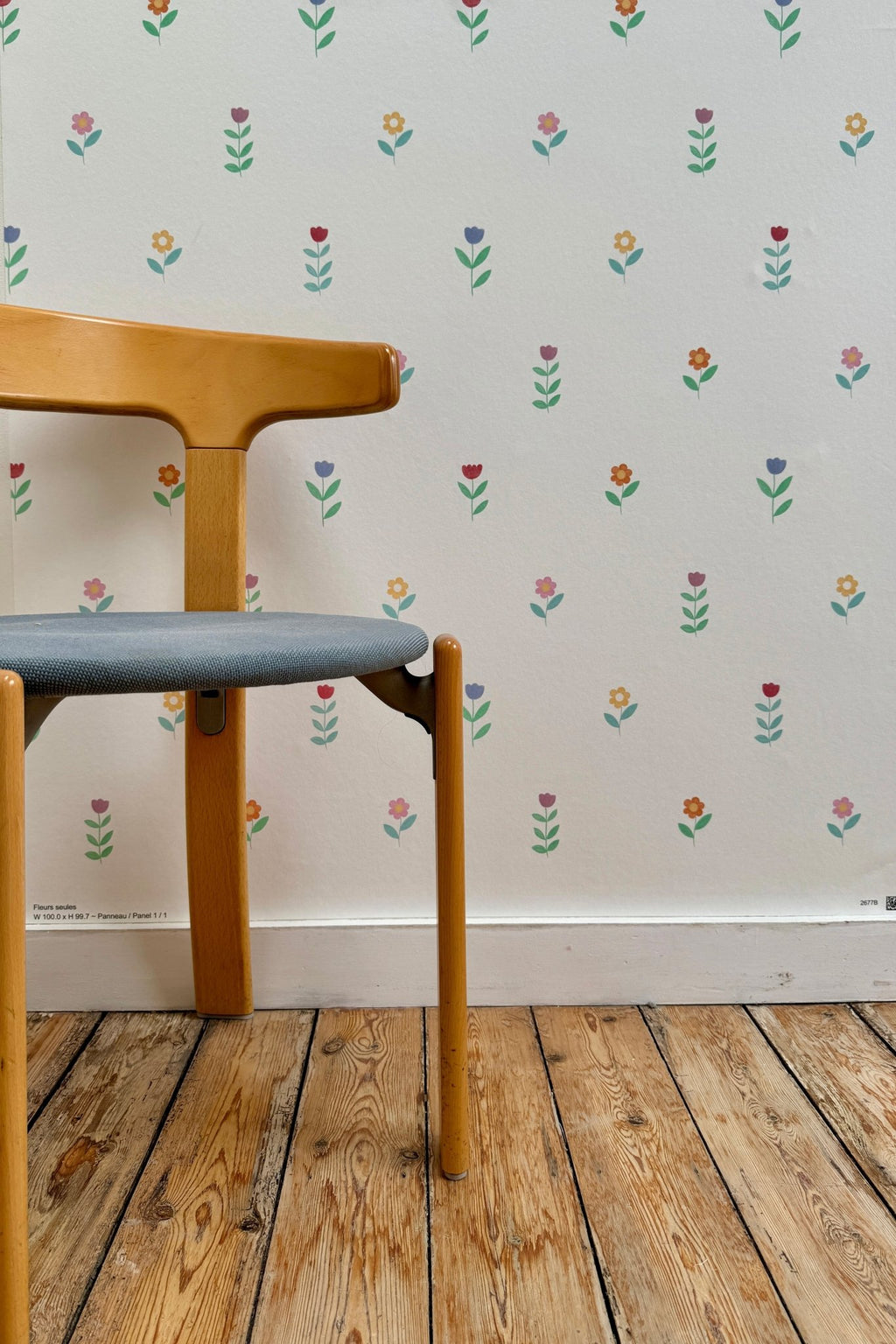 Échantillon du papier peint Juliette à motif de petites fleurs colorées, tapissé sur un mur derrière une chaise en bois vintage à assise grise posée sur un parquet brut.