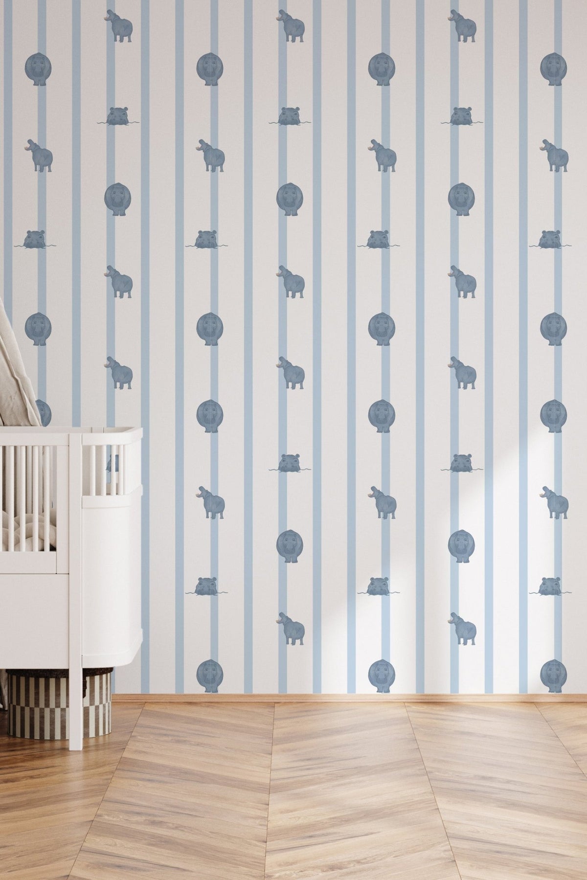 Échantillon du papier peint Paul à rayures bleues et motifs hippopotames, décorant une chambre de bébé lumineuse avec un lit à barreaux blanc posé sur un parquet en chevron.