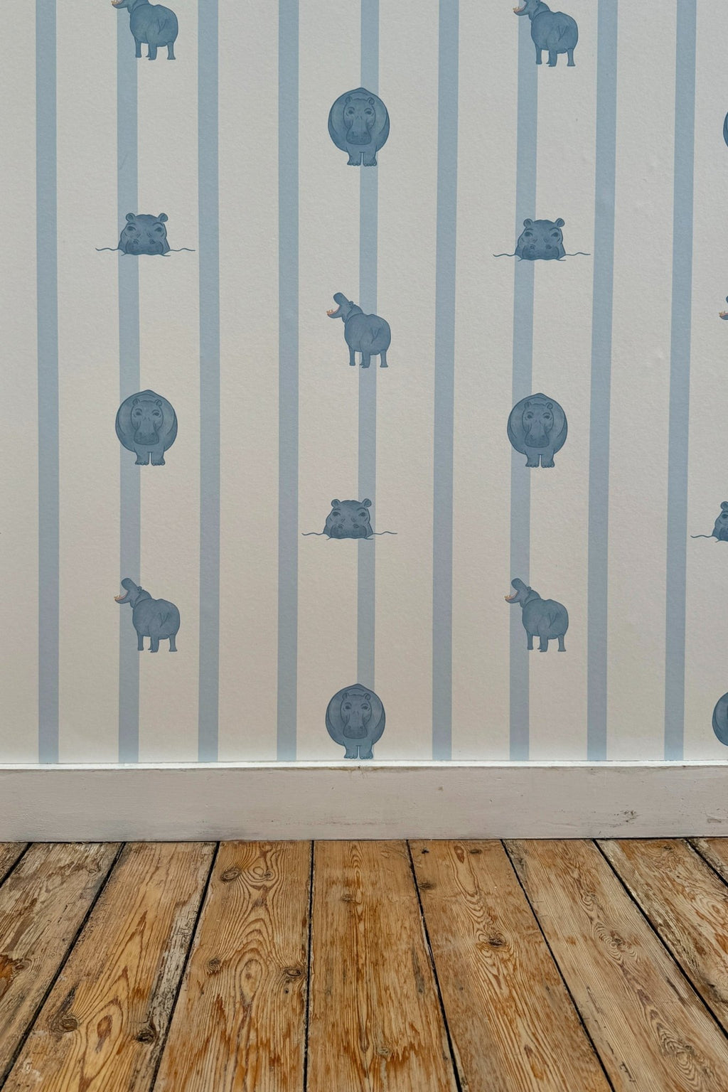 Échantillon du papier peint Paul à motif ligné bleu clair et petites illustrations d'hippopotames, posé sur un mur au-dessus d'une plinthe blanche et d'un parquet en bois brut.