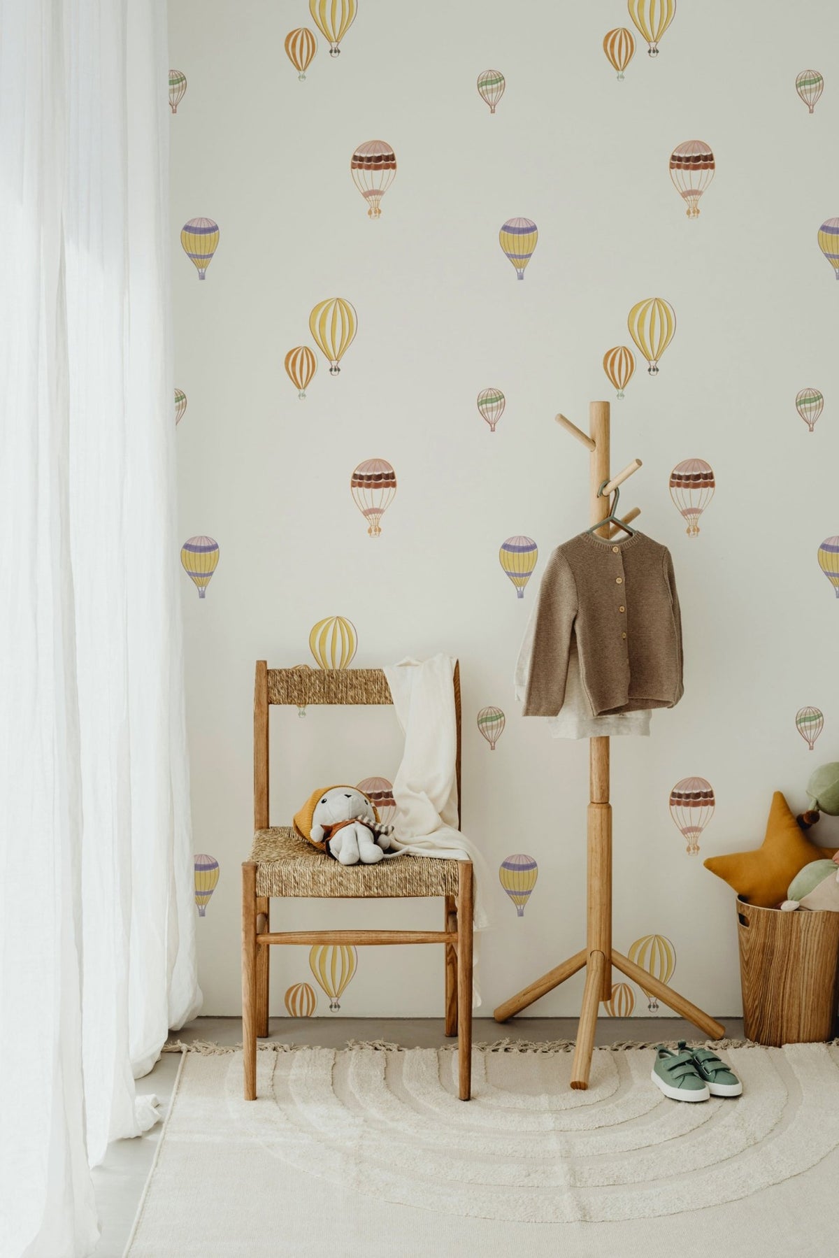 Échantillon du papier peint Edgard à motif de montgolfières colorées, derrière une chaise en bois tressé et un porte-manteau d'enfant.