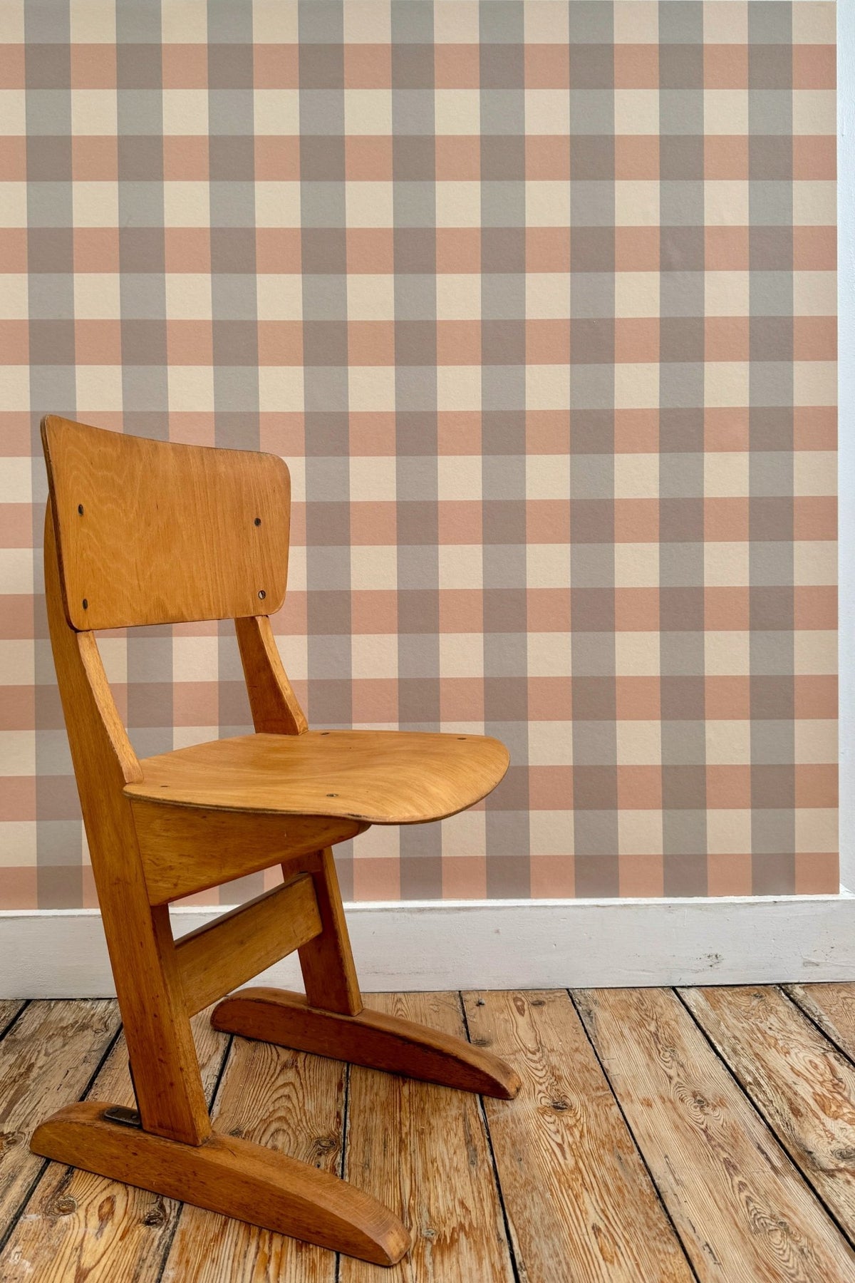 Échantillon du papier peint Karl à motif vichy gris et marron, tapissé sur un mur derrière une chaise d'écolier vintage en bois posée sur un parquet brut.