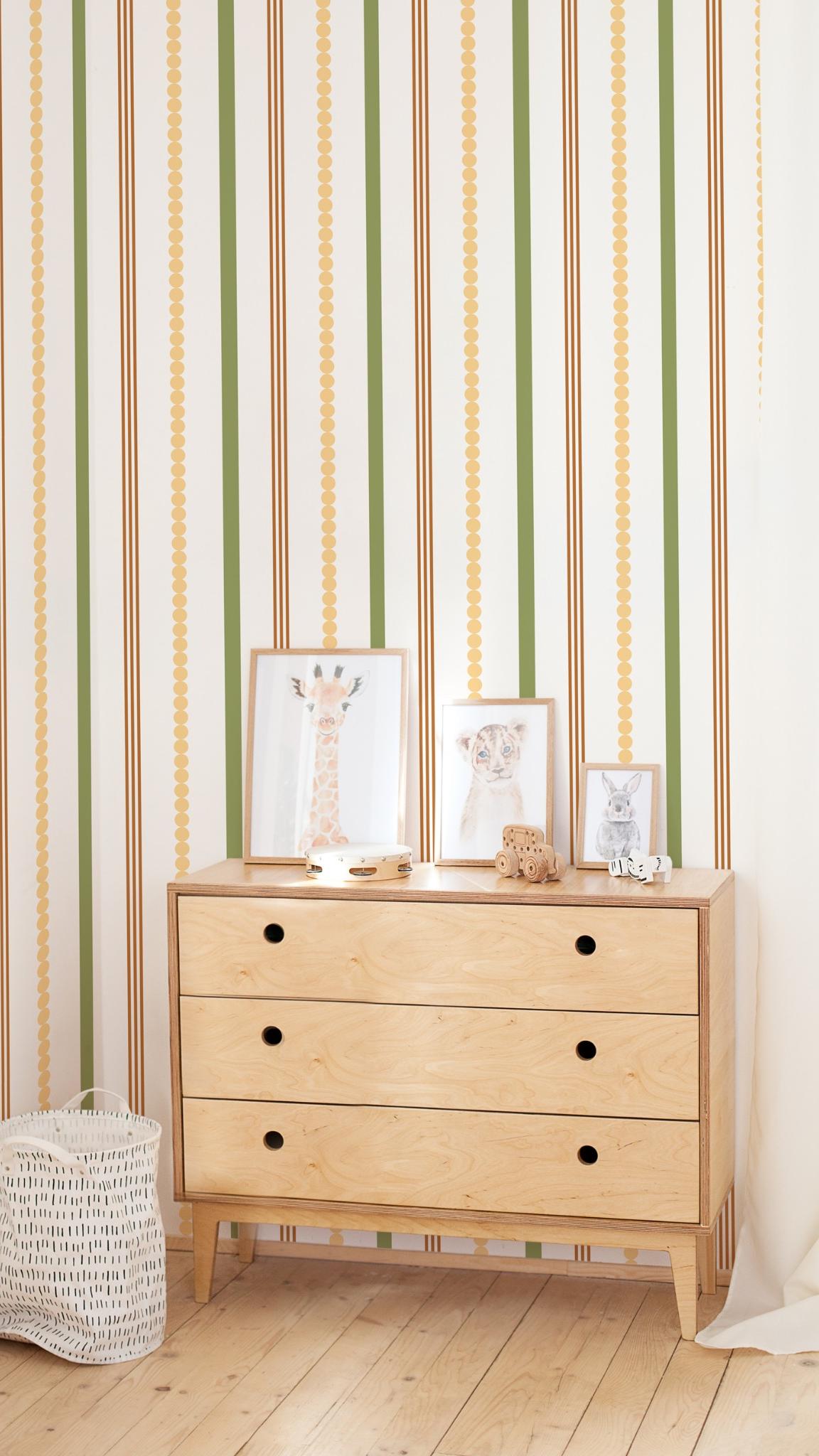 Papier peint Amina à rayures verticales vertes, marron et points jaunes, posé derrière une commode en bois dans une chambre d'enfant.