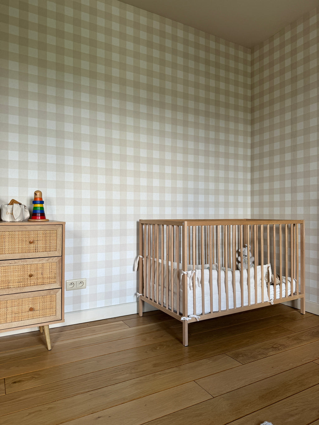 Papier peint Ernest à carreaux vichy beige et blanc tapissé sur les murs d'une chambre de bébé, avec un lit à barreaux en bois et une petite commode en rotin.