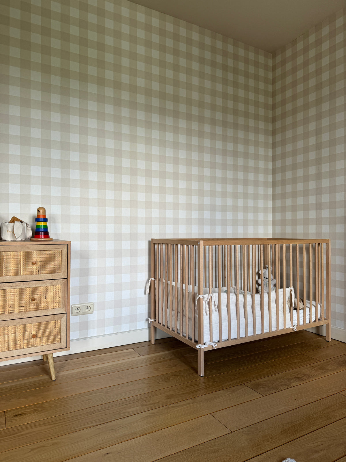 Papier peint Ernest à carreaux vichy beige et blanc tapissé sur les murs d'une chambre de bébé, avec un lit à barreaux en bois et une petite commode en rotin.