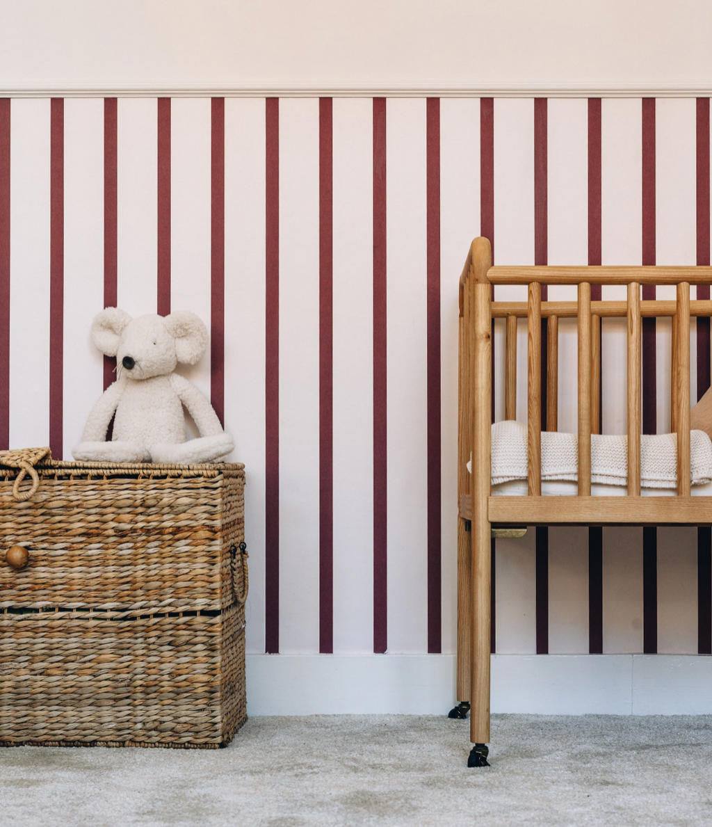 Papier peint Clarisse à larges rayures cerise sur fond rose pâle, décorant une chambre de bébé avec un berceau en bois et un panier en osier.