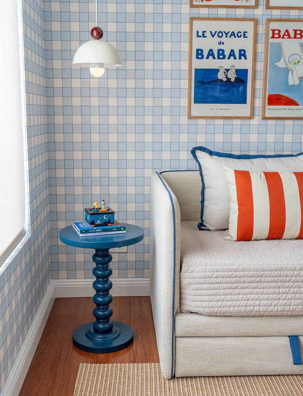 Papier peint Jack à motif damier bleu et blanc, décorant un coin nuit pour enfant avec un lit banquette, un coussin rayé rouge, une petite table bleue sculptée et des affiches vintage encadrées.