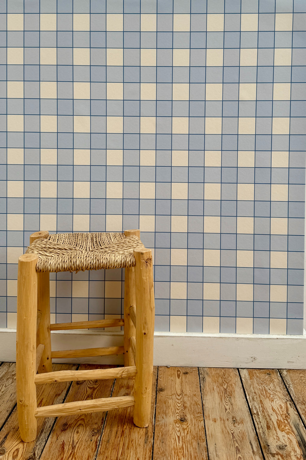 Papier peint Jack à motif damier bleu et blanc, posé sur un mur derrière un tabouret rustique en bois à assise tressée posé sur un parquet brut.