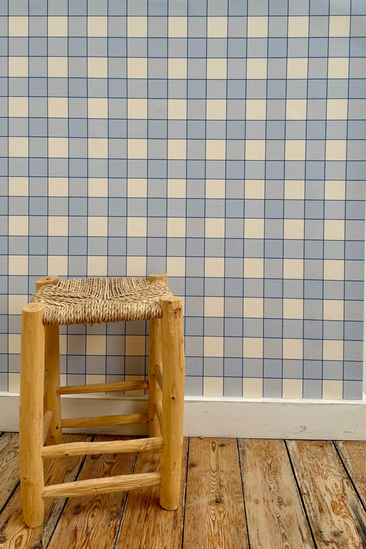 Papier peint Jack à motif damier bleu et blanc, posé sur un mur derrière un tabouret rustique en bois à assise tressée posé sur un parquet brut.