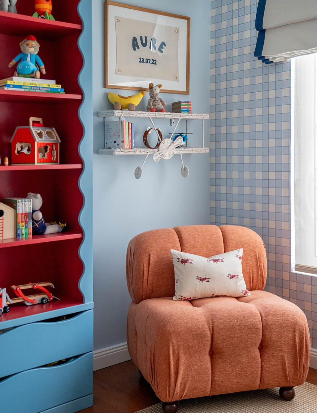 Papier peint Jack à motif damier bleu et blanc, décorant un coin lecture pour enfant avec un fauteuil capitonné orange, une étagère murale en forme d'avion et une grande bibliothèque rouge remplie de jouets.