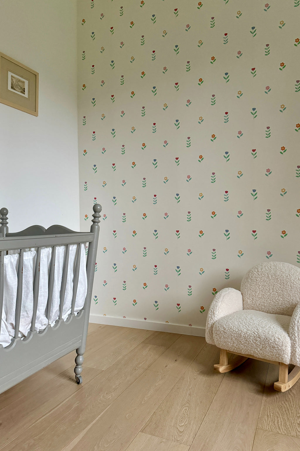 Papier peint Juliette à motif de petites fleurs, décorant une chambre de bébé avec un lit à barreaux gris vintage et un petit fauteuil à bascule en tissu bouclette blanc.