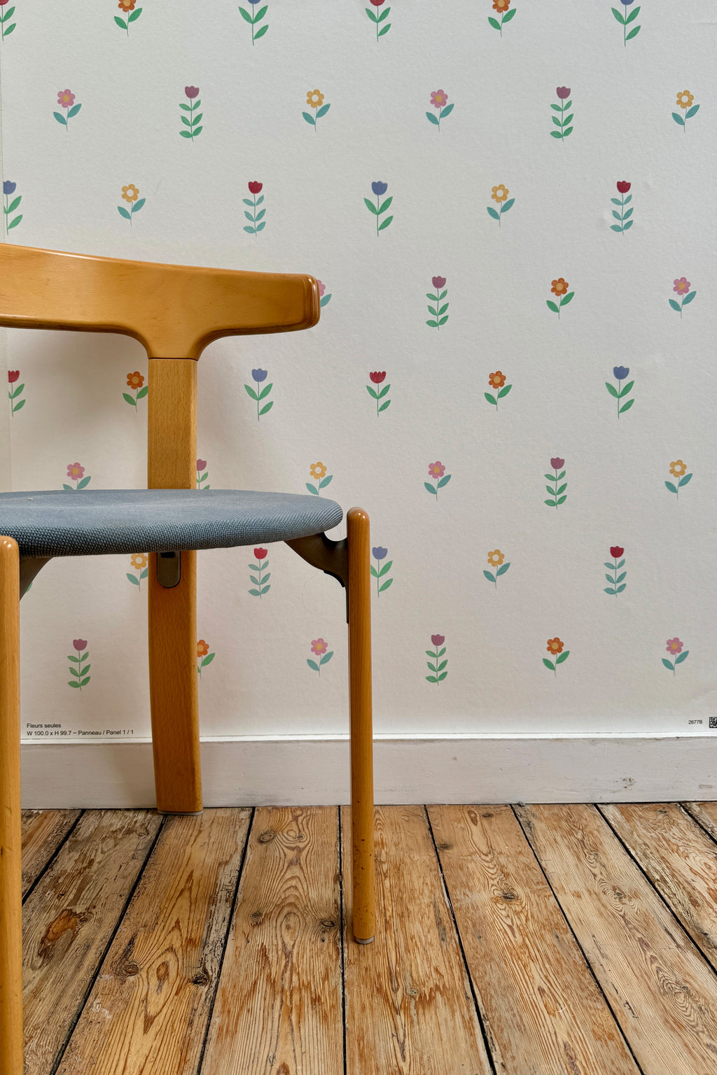 Papier peint Juliette à motif de petites fleurs colorées, tapissé sur un mur derrière une chaise en bois vintage à assise grise posée sur un parquet brut.