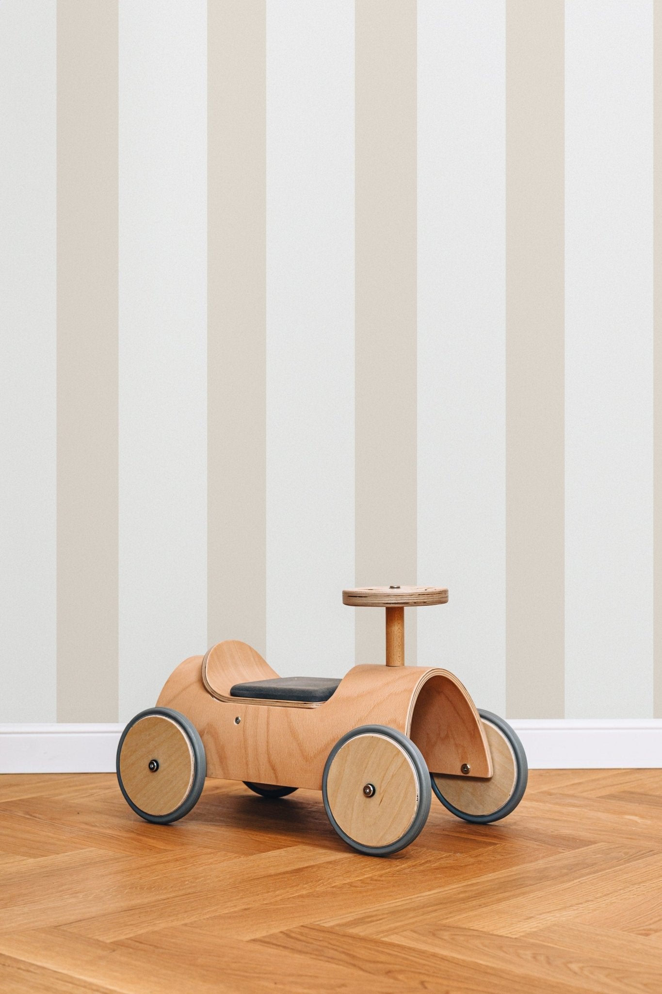 Papier peint Jules à larges rayures beiges, posé dans une chambre d'enfant avec un porteur en bois sur un parquet.