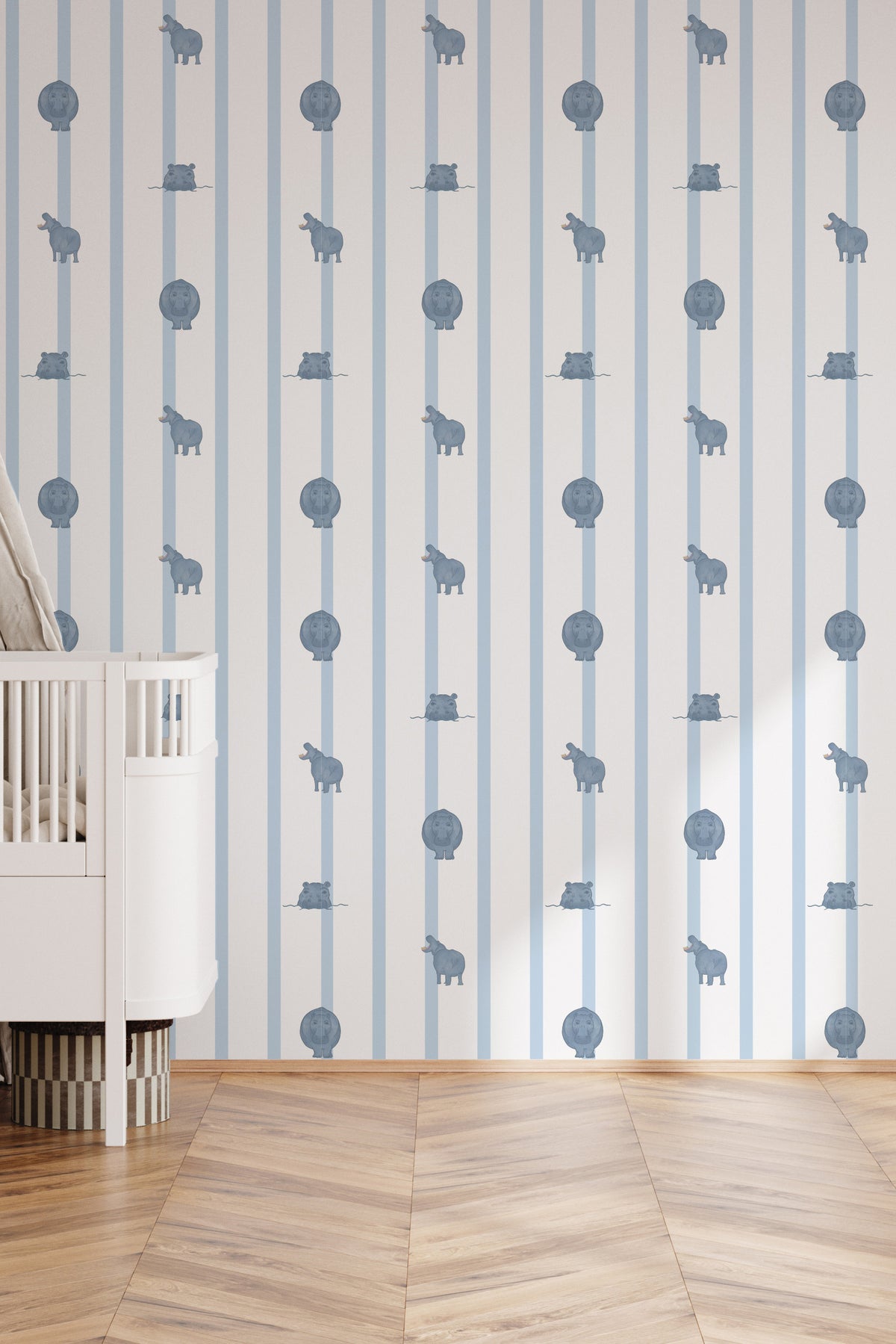 Papier peint Paul à rayures bleues et motifs hippopotames, décorant une chambre de bébé lumineuse avec un lit à barreaux blanc posé sur un parquet en chevron.