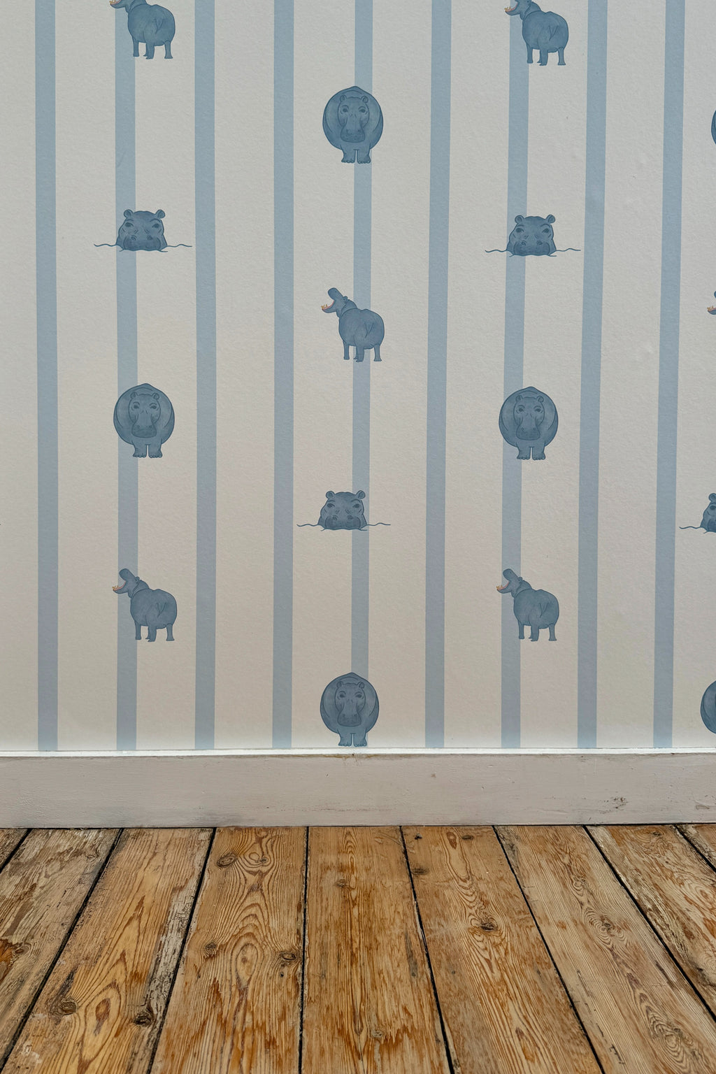 Papier peint Paul à motif ligné bleu clair et petites illustrations d'hippopotames, posé sur un mur au-dessus d'une plinthe blanche et d'un parquet en bois brut.