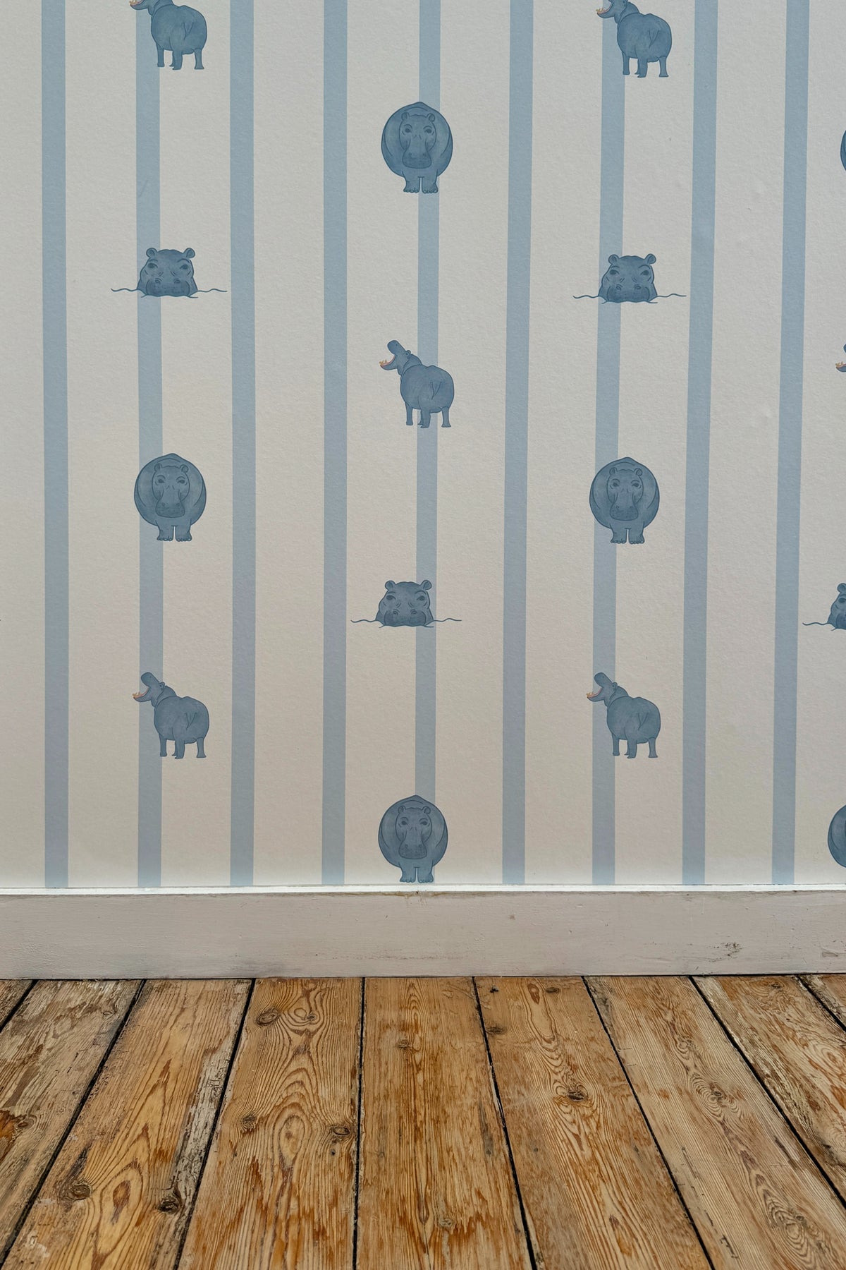 Papier peint Paul à motif ligné bleu clair et petites illustrations d'hippopotames, posé sur un mur au-dessus d'une plinthe blanche et d'un parquet en bois brut.