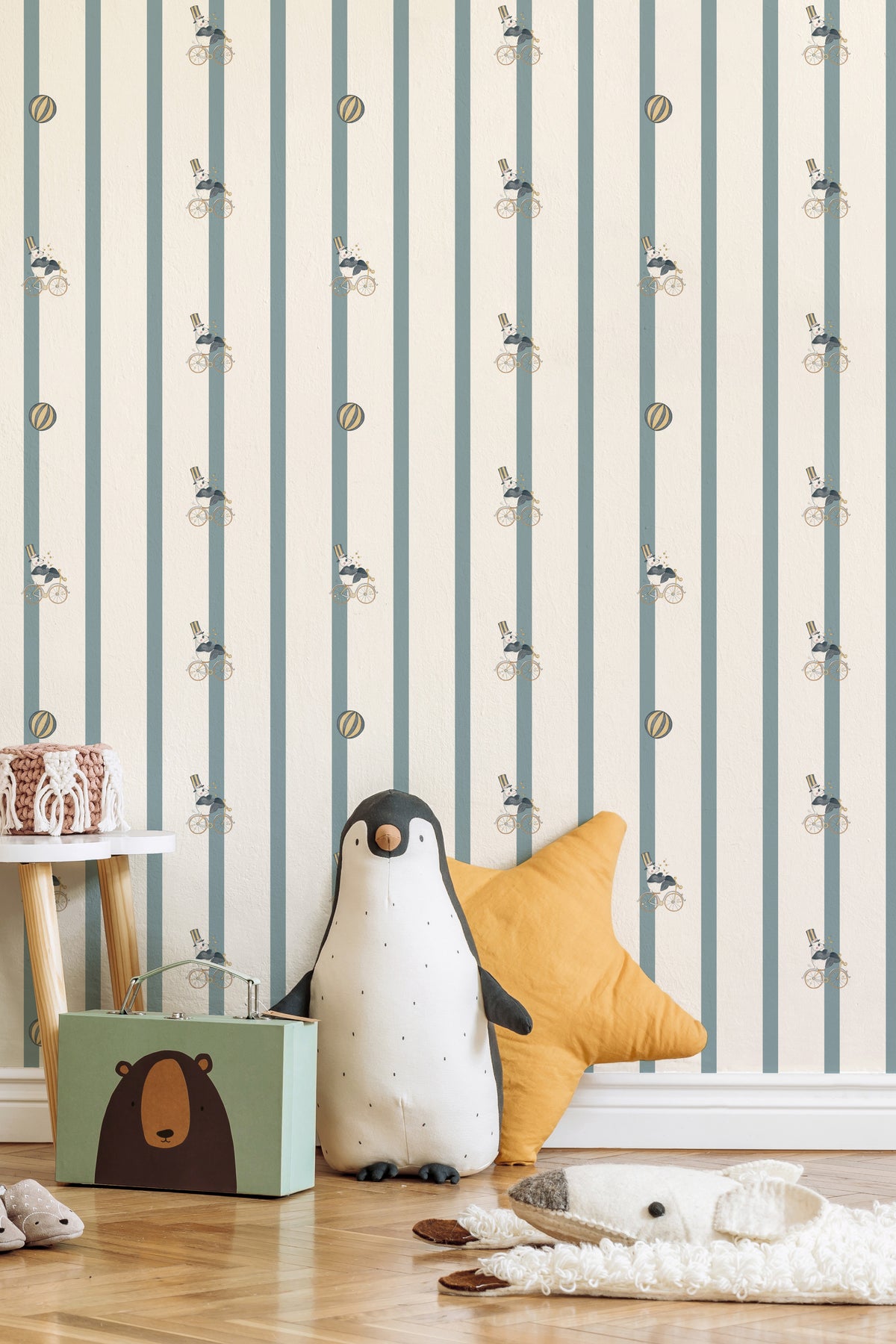 Papier peint Nelson ligné bleu gris et blanc avec motifs de pandas sur des vélos jaunes, mis en scène dans une chambre d'enfant ludique avec une grande peluche pingouin, un coussin étoile jaune et un petit tabouret en bois.