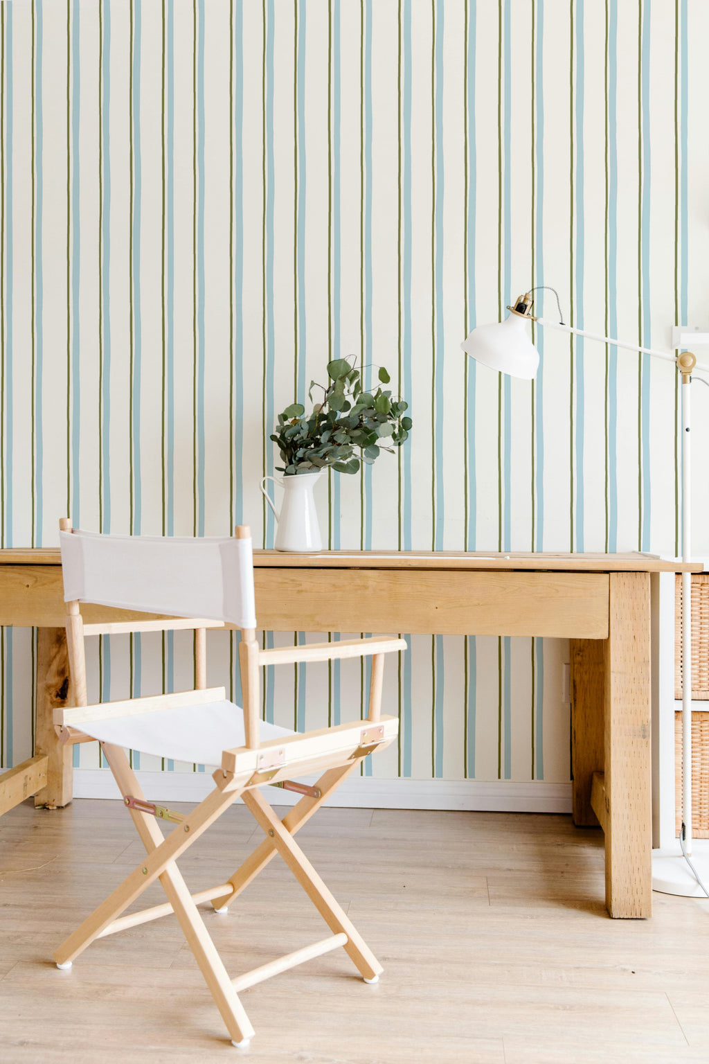Papier peint Celeste à motif ligné bleu et vert, décorant un espace bureau lumineux avec une grande table en bois clair, une chaise de metteur en scène en toile blanche et un vase avec de l'eucalyptus.