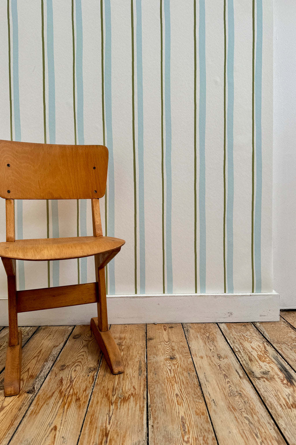 Papier peint Celeste à motif ligné bleu et vert, posé sur un mur derrière une chaise d'écolier vintage en bois sur un parquet brut.