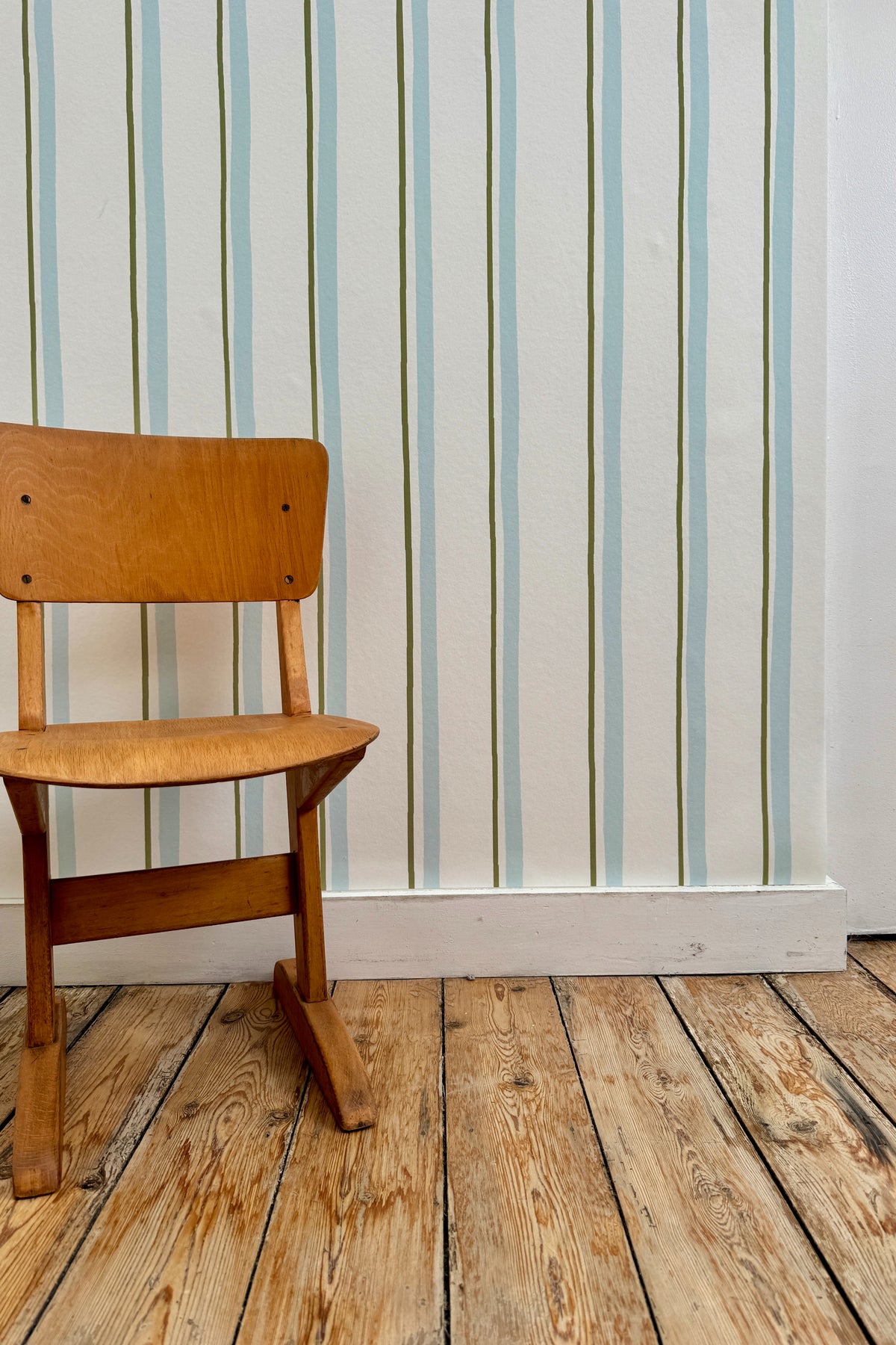 Papier peint Celeste à motif ligné bleu et vert, posé sur un mur derrière une chaise d'écolier vintage en bois sur un parquet brut.