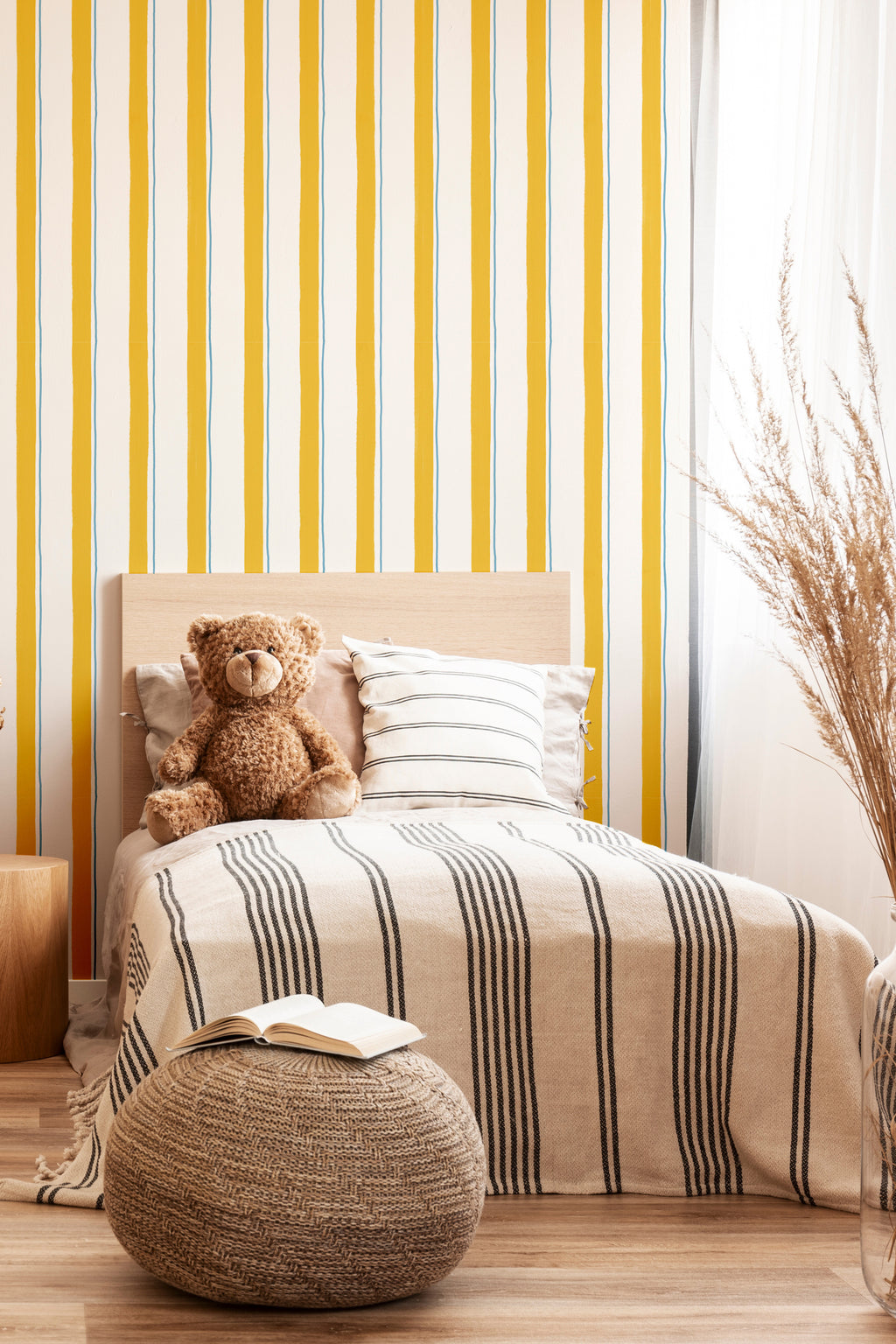 Papier peint Victor à larges lignes jaunes et fines lignes bleues claires, décorant une chambre d'enfant chaleureuse avec un lit en bois, un ours en peluche, un grand pouf tressé et un vase avec des herbes de la pampa.