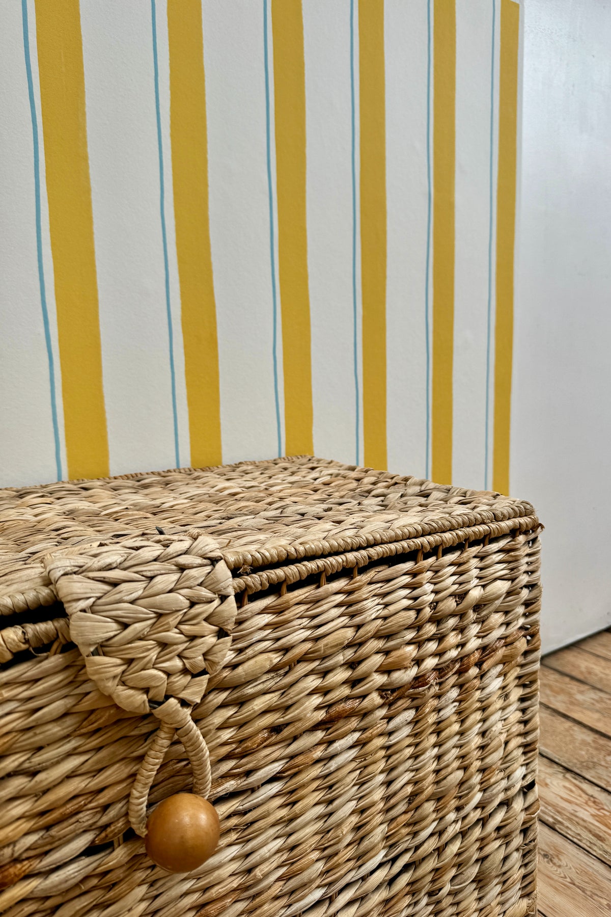 Papier peint Victor avec de larges lignes jaunes et de fines lignes bleues claires, posé sur un mur juste derrière une grande malle de rangement en osier tressé sur un parquet.