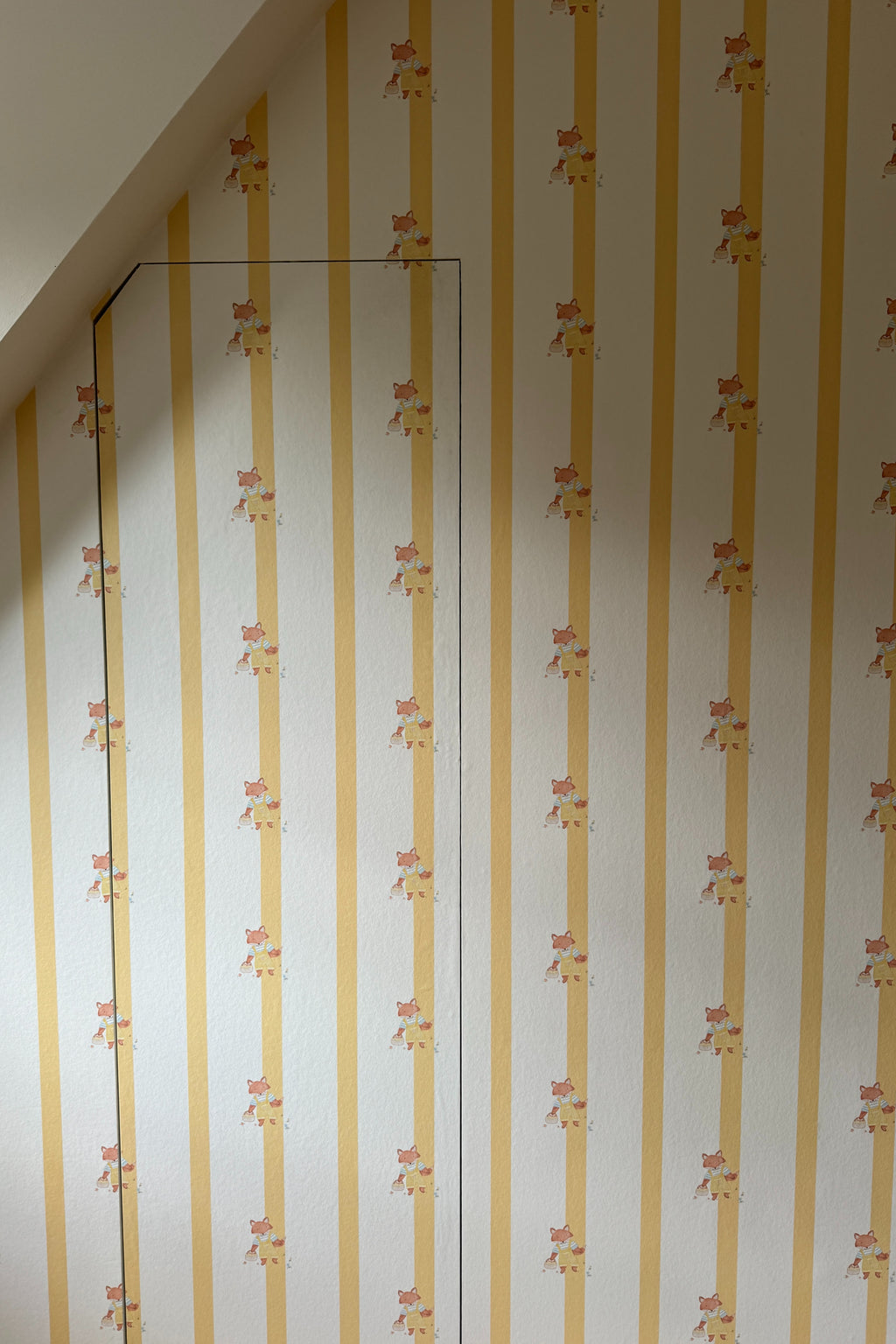 Papier peint Joy à rayures jaunes et petits motifs de renards, tapissé sur un mur en sous-pente avec une porte invisible parfaitement raccordée au motif.