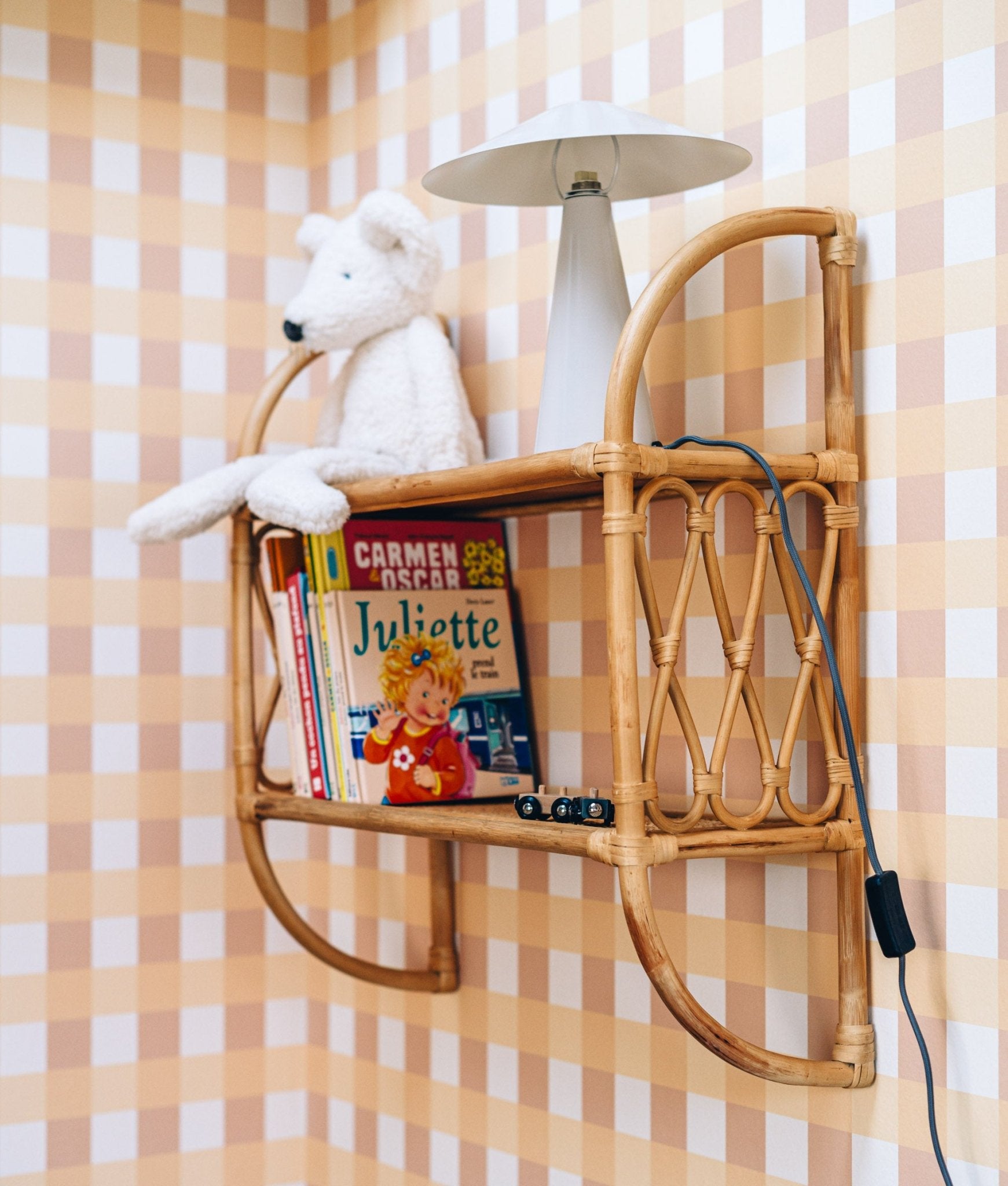Papier peint Lola à motif vichy camel, posé derrière une étagère en rotin avec des livres et une peluche.