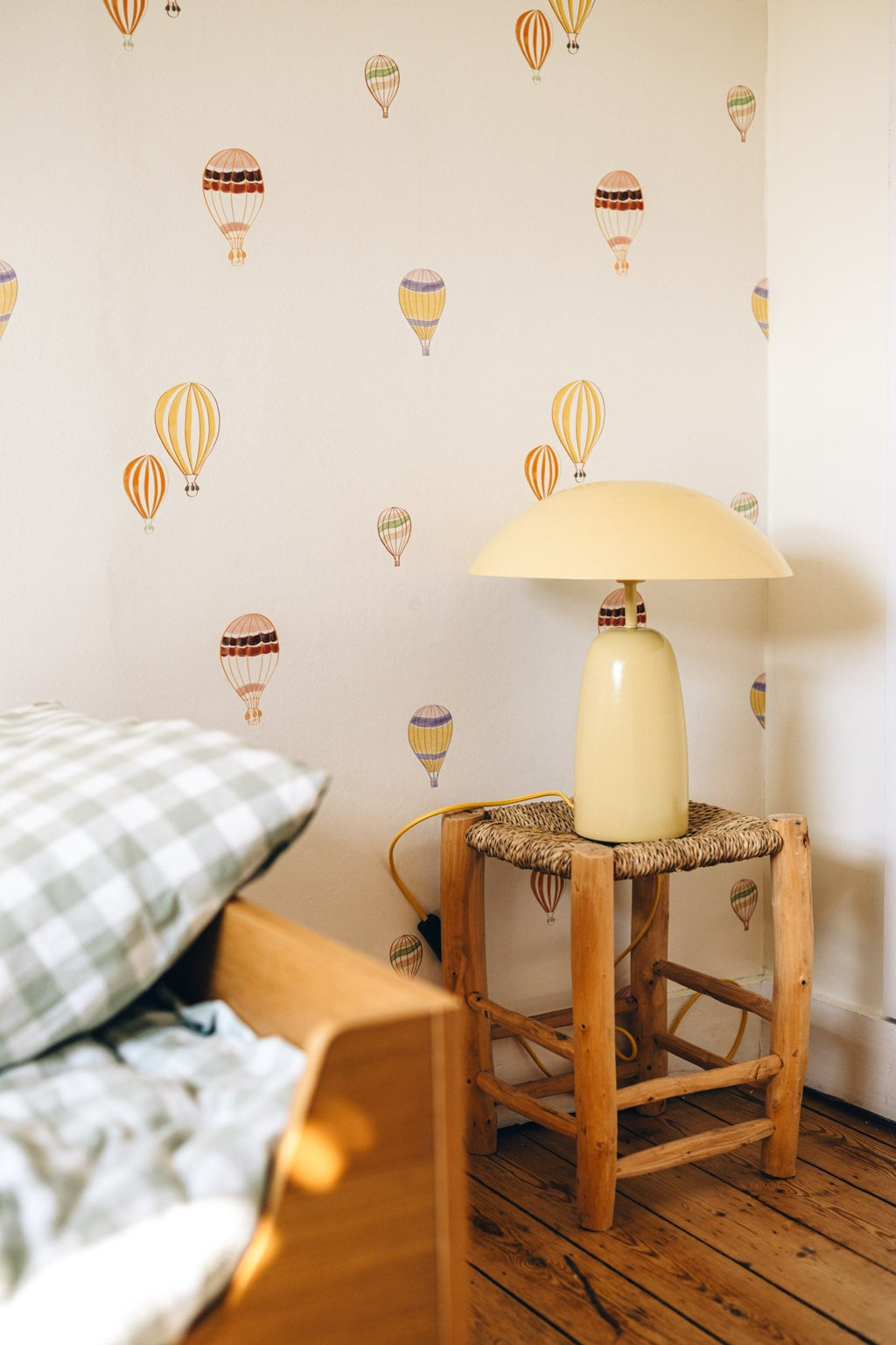 Papier peint Edgard à motif montgolfières, au-dessus d'un lit en bois et d'un tabouret rustique avec une lampe jaune.