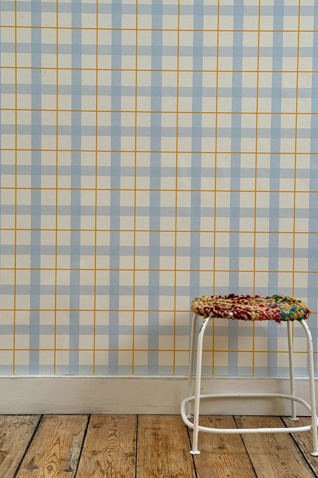 Papier peint Auguste à motif quadrillé bleu et jaune, posé sur un mur au-dessus d'un parquet en bois brut avec un petit tabouret tressé coloré.