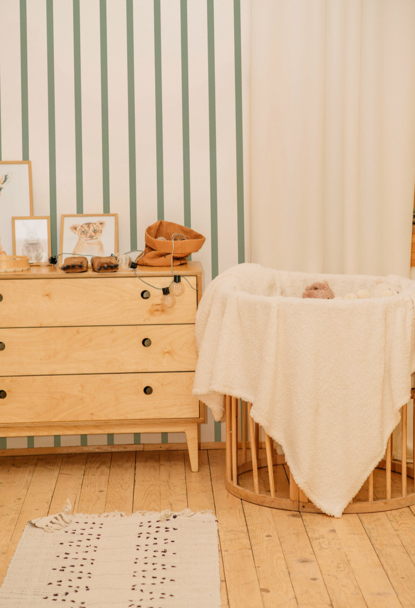 Papier peint Michèle à rayures verticales vert sauge et crème, mis en situation dans une chambre de bébé douce avec une commode en bois clair et un berceau.