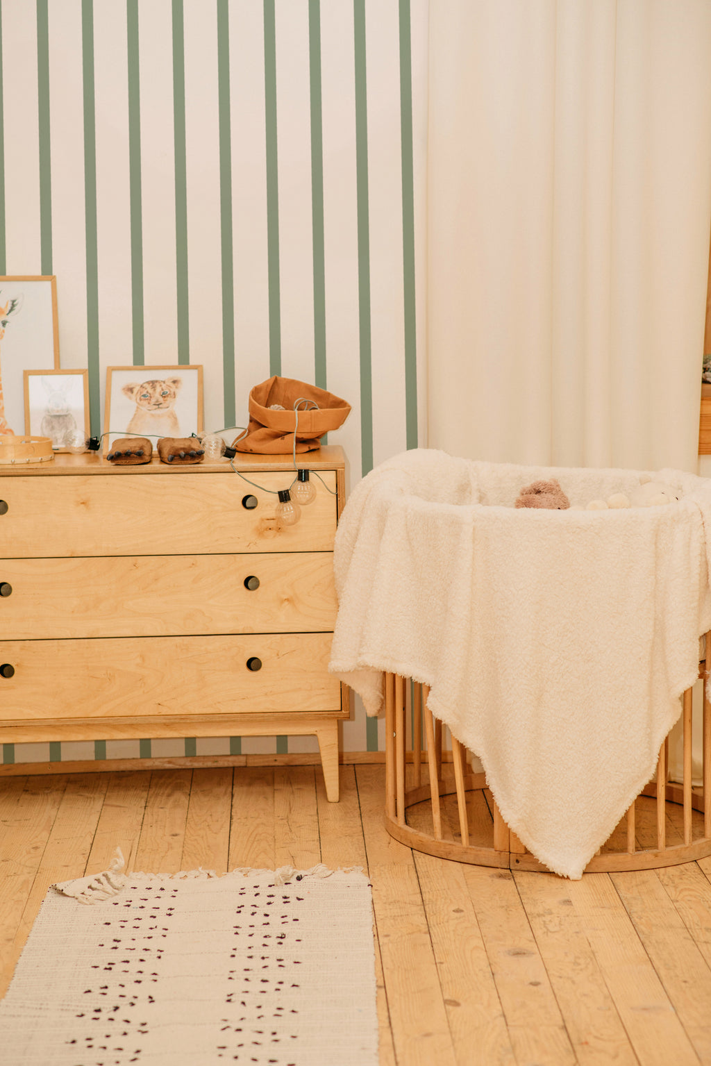 Papier peint Michèle à rayures vertes sauge et beige, derrière une commode en bois et un berceau recouvert d'un plaid blanc.