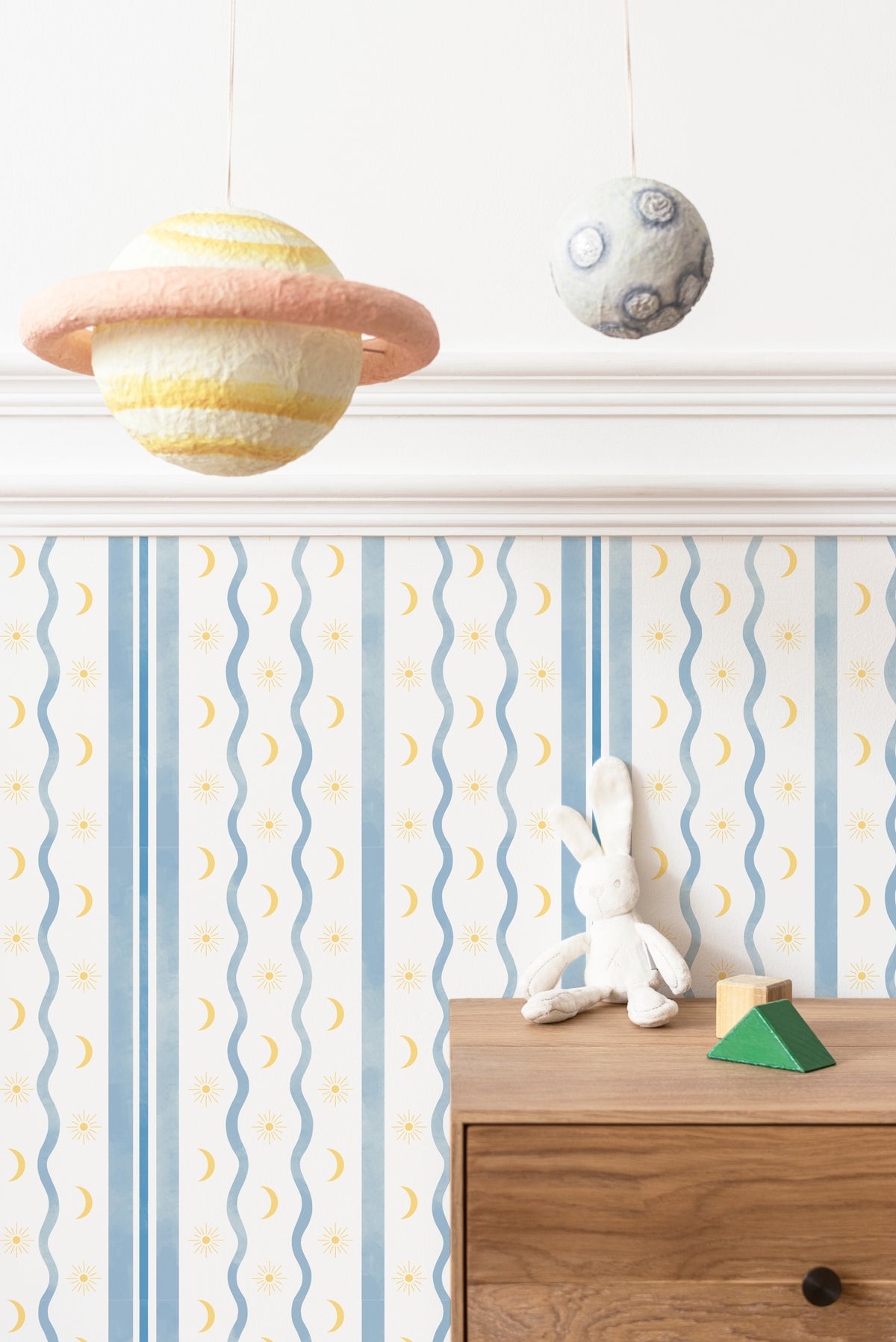Papier peint enfant à rayures bleues, lunes et soleils, au-dessus d'une commode en bois avec une peluche lapin.