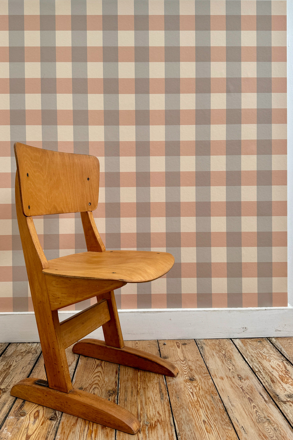 Papier peint Karl à motif vichy gris et marron, tapissé sur un mur derrière une chaise d'écolier vintage en bois posée sur un parquet brut.