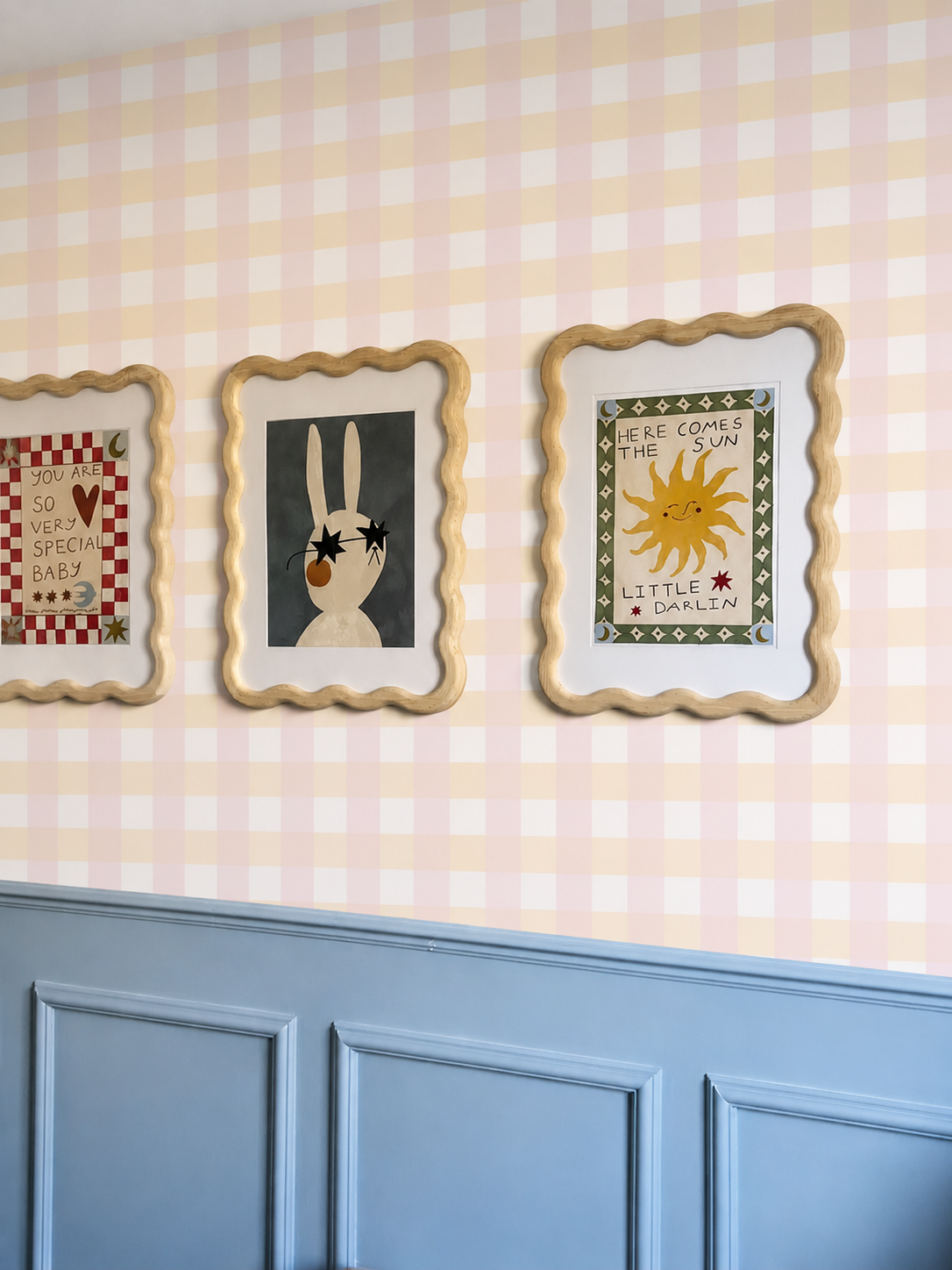 Papier peint à carreaux vichy pastel Dalia posé au-dessus d'un soubassement bleu avec des cadres décoratifs muraux.