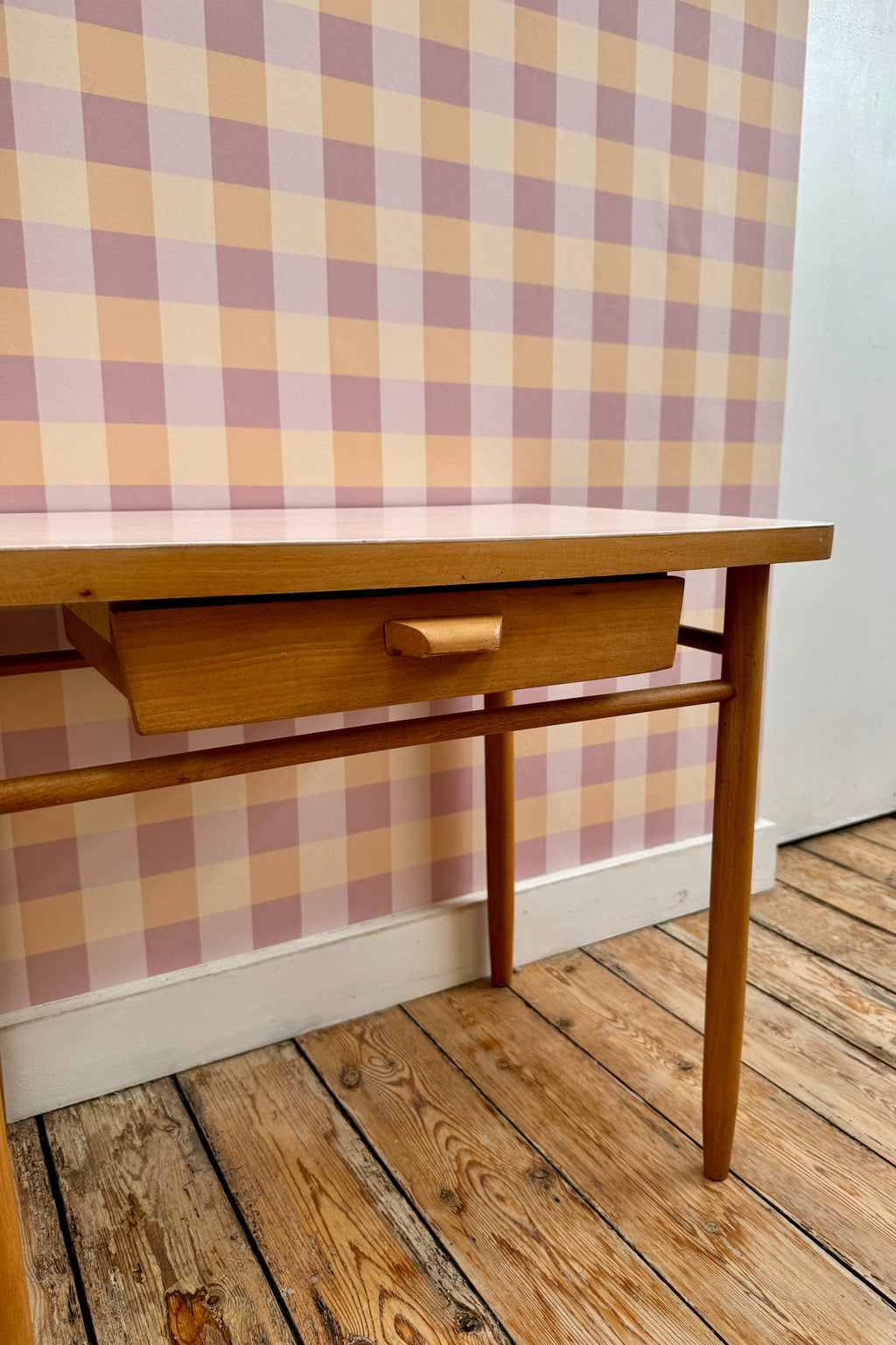 Papier peint Cerise à motif vichy rose et orange, posé sur un mur derrière un petit bureau d'écolier vintage en bois.