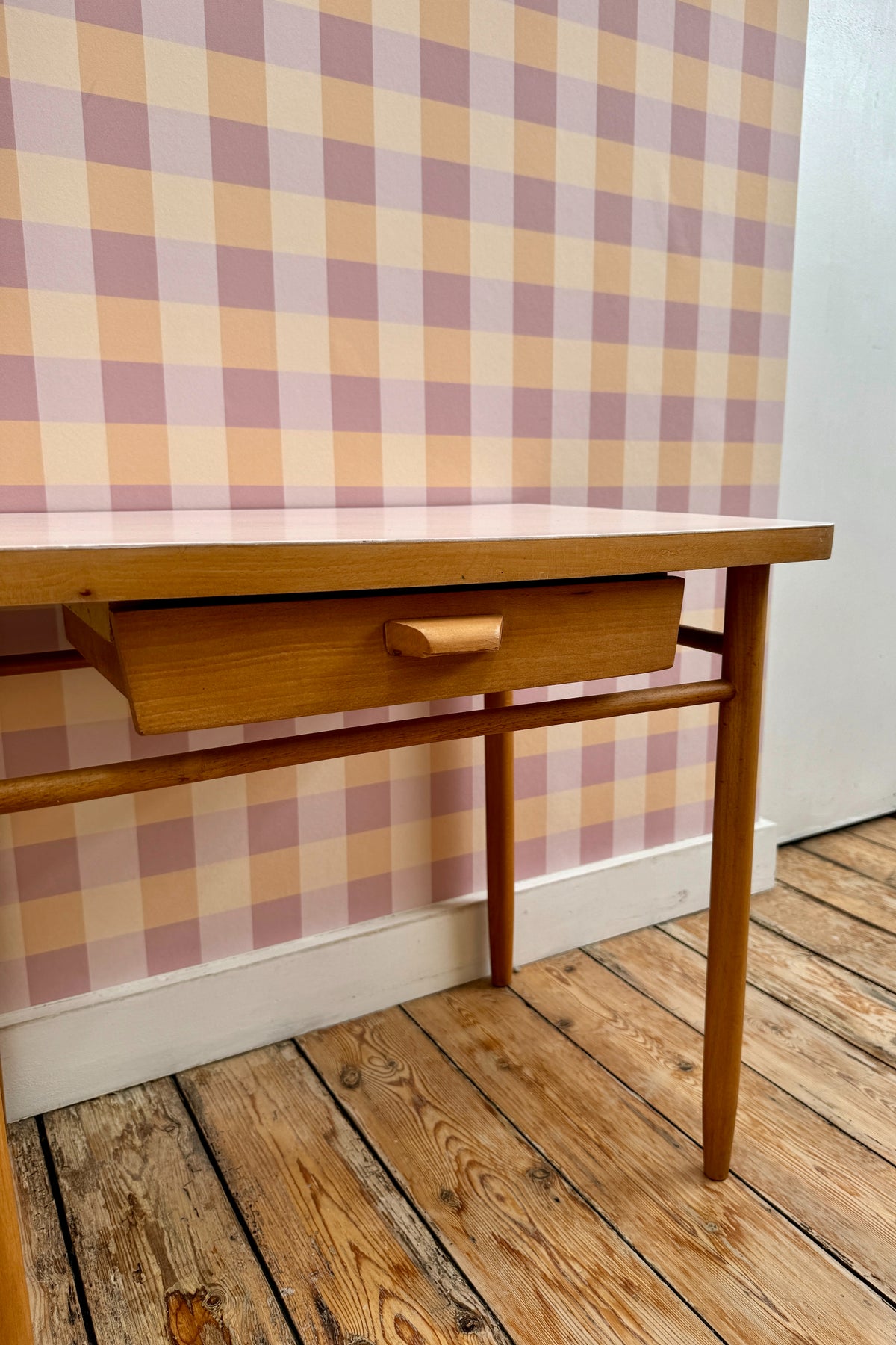 Papier peint Cerise à motif vichy rose et orange, posé sur un mur derrière un petit bureau d'écolier vintage en bois.