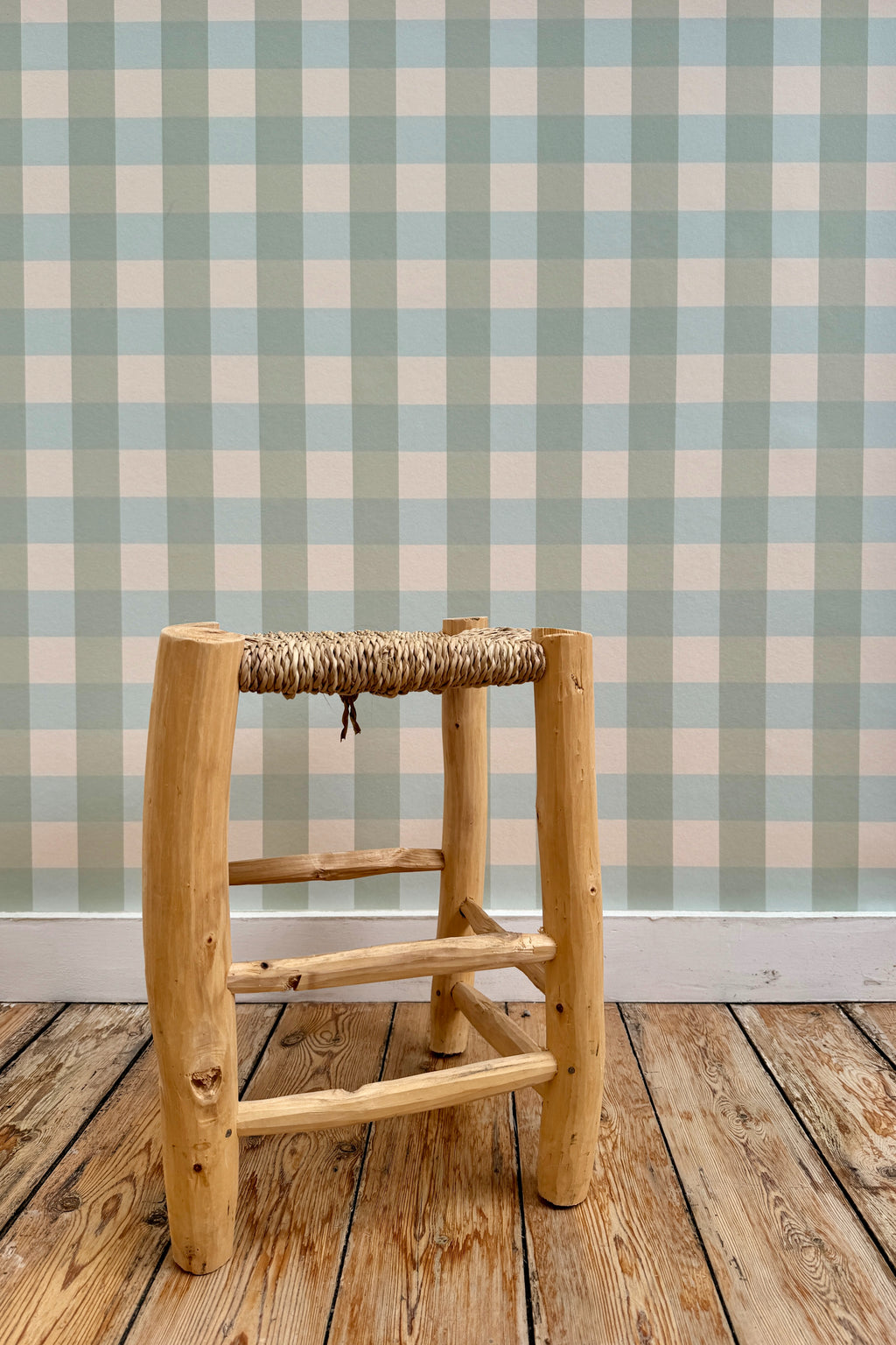 Papier peint Brieuc à motif vichy vert et bleu, tapissé sur un mur derrière un tabouret rustique en bois à assise tressée posé sur un parquet brut.
