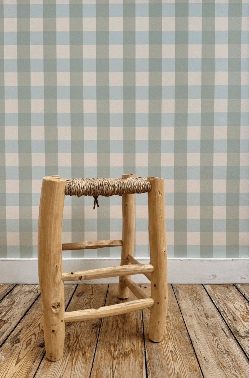 Papier peint Brieuc à motif vichy vert et bleu, tapissé sur un mur derrière un tabouret rustique en bois à assise tressée posé sur un parquet brut.